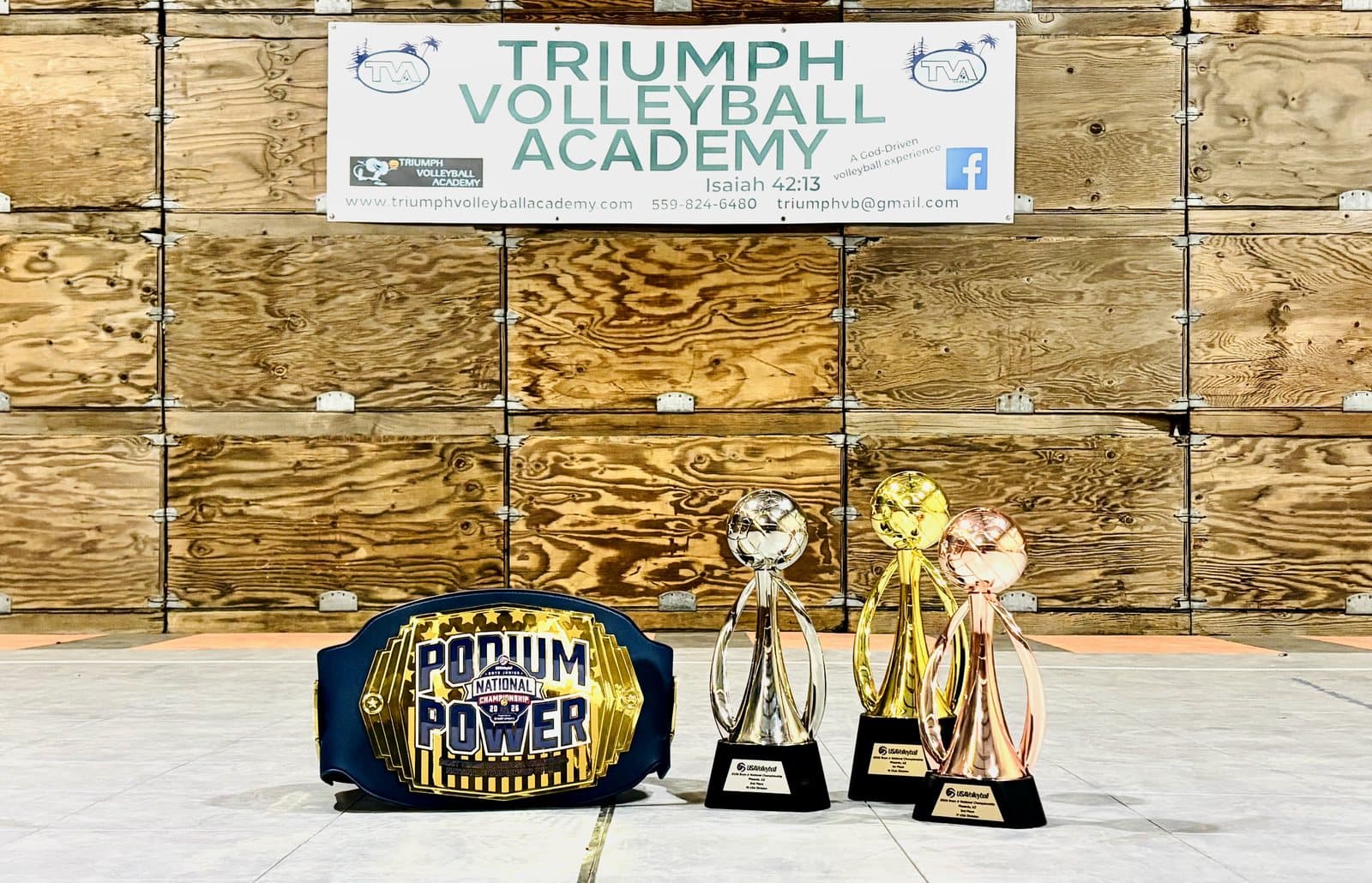 Podium Power championship belt and the gold, silver and bronze trophies TVA boys won at the 2026 USA Volleyball National Championships in Phoenix, displayed under the Triumph Volleyball Academy banner